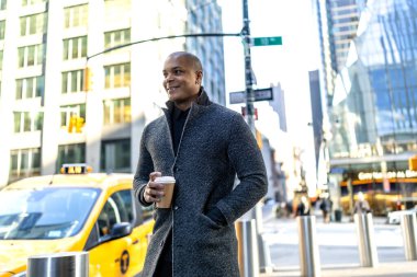 Kendine güvenen siyah profesyonel şık ceketli, hazır kahve fincanı tutan, kalabalık bir Manhattan caddesinde sarı taksi ve modern binalarla yürüyen, kentsel iş günü enerjisini somutlaştıran