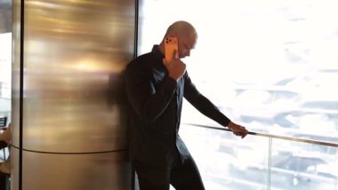 Metalik bir duvara yaslanmış, akıllı telefonuyla konuşan, iş ilişkilerini büyük bir pencereden parlak, güneşli şehir manzarasıyla yöneten, kendinden emin genç bir işadamı.