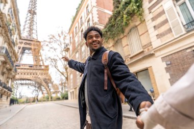 Afrika kökenli mutlu genç bir adam Paris, Fransa 'da romantik bir şehir molası sırasında gülümserken Eyfel Kulesi' ni işaret ederek ortağının elinden tutar.