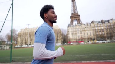 Kararlı Afrikalı Amerikalı sporcu günlük kardiyo antrenmanının keyfini çıkarıyor, güzel Paris arka planında görünen ikonik Eyfel kulesiyle bir stadyumda koşuyor.
