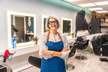Kıdemli kadın kuaför gülümsüyor, kolları çapraz duruyor, gözlük takıyor ve önlük takıyor, kuaför dükkanını güvenle yönetiyor, girişimciliği ve küçük işletme başarısını temsil ediyor.