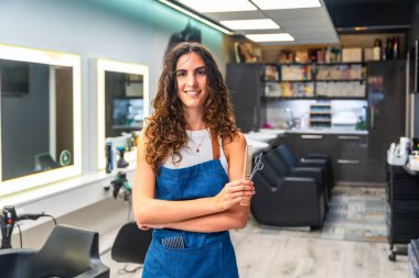 Kadın kuaför gülümsüyor, küçük bir kuaförde duruyor, elinde saç makasıyla tarak tutuyor, profesyonelliği, uzmanlığı ve güzellik ve stile olan bağlılığı simgeliyor.