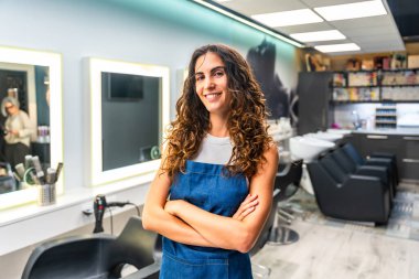 Kıvırcık saçlı, önlük takan ve gülümseyen, modern kuaföründe kollarını kavuşturmuş, küçük bir işletme sahibini temsil eden kendine güvenen genç bir kadın.
