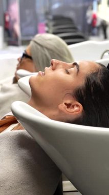 Rahatlamış genç bir kadın saçını yıkayıp duruluyor. Modern bir güzellik salonundaki bir kuaför tarafından. Profesyonel saç bakımının keyfini çıkarırken gülümsüyor.
