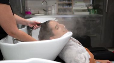 Modern bir güzellik salonunda profesyonel bir saç buharı ve yıkama tedavisinin keyfini çıkaran rahat genç bir kadın. Lavaboda kafa derisine masaj yapan bir kuaför var.