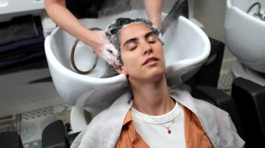 Gözleri kapalı genç bir kadın, modern bir güzellik salonundaki profesyonel bir kuaförden kafa masajı ve buharlı saç yıkama tedavisiyle keyif alıyor. Sağlık ve kendine bakımı teşvik ediyor.