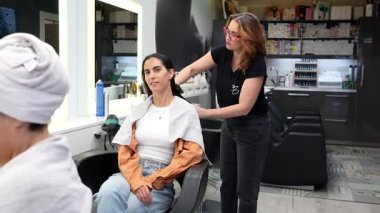 Kadın kuaför, müşterilerinin ıslak saçlarını tararken aynanın önünde bir sandalyede oturuyor, profesyonel ve modern bir güzellik salonunda yeni bir saç kesimi ve stil oturumuna hazırlanıyor.