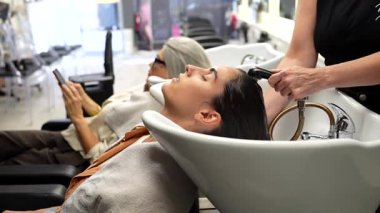 Profesyonel bir kuaför, kuaförde genç bir kadının saçını yıkayarak rahatlatıcı bir şampuan ve kondisyon tedavisi sunuyor. Bu sırada da diğer müşteri arka planda bekleyip gülüyor.