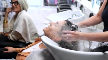 Modern bir güzellik salonunda profesyonel kafa masajının ve buharla yıkanan saçların keyfini çıkaran rahat genç bir adam, diğer bir müşteri berber dükkanının arka planında bekliyor ve konuşuyor.