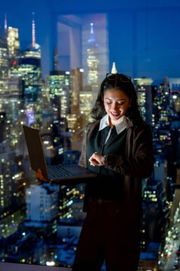 Geniş bir pencerenin yanında duran genç iş kadını, bir dizüstü bilgisayarla çalışıyor ve şehir ışıkları, özveri ve başarıyı temsil eden arka planı aydınlatırken, akıllı saatlerini kontrol ediyor.
