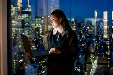 Genç iş kadını, elinde bir dizüstü bilgisayarla elinde bir kattan tavana kadar ayakta duruyor ve gökyüzünü aydınlatan Manhattan 'a gülümsüyor. Hırsı, liderliği ve modern çalışma hayatını simgeliyor.