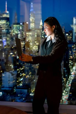 Genç iş kadını yüksek bir pencerenin önünde duruyor, aktif olarak dizüstü bilgisayarla çalışıyor, Manhattan şehrinin gökdelenleri ve gökdelenleri gece görülebiliyor.