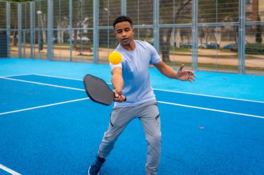 Genç adam, canlı mavi bir sahada raketle sarı bir picleball topuna vuruyor. Modern eğlence sporlarında enerjik hareketler, odaklanma ve aktif yaz tatili eğlencesi sergiliyor.