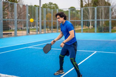 Genç bir adam mavi bir açık hava sahasında Pickleball oynuyor, siyah bir kürekle sarı bir topa vuruyor, atletik hareketler gösteriyor, odaklanıyor ve spor giyim tarzında aktif bir eğlence yaşam tarzı.