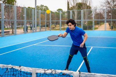 Mavi takım elbiseli genç adam mavi bir kortta, dikkatle sarı topa kürekle vuruyor, modern raket sporunun tadını çıkarıyor.
