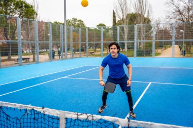 Genç bir adam canlı mavi bir sahada, bir raket tutuyor ve maç sırasında topu izliyor. Sağlıklı ve aktif bir yaşam tarzını canlandırıyor.
