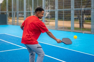 Spor giyim sektöründe genç bir atletik adam canlı mavi bir sahada raketle pena topuna vuruyor, odaklanıp gülümsüyor ve enerjik yaz oyunları ve formdan keyif alıyor.