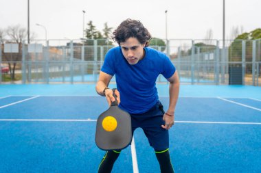 Spor giyimli genç bir adam servise konsantre oluyor. Pickleball topunu kavrıyor. Sarı top açık hava sahasının üzerinde süzülüyor.