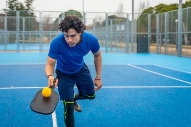 Erkek sporcu, elinde bir picleball raketi ve sarı bir top tutarken konsantre oluyor. Canlı bir mavi sahada oynamaya ya da dönmeye hazır. Aktif spor ve formda.