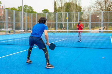 İki erkek sporcu, canlı bir mavi sahada, güçlü bir picleball oyunu oynuyorlar, raketleri tutuyorlar ve açık havada ve aktif bir spor müsabakası sırasında topa vurmaya odaklanıyorlar.