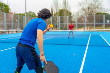 Pickleball oyuncusu mavi bir sahada bir rakiple karşılaşmaya hazırlanıyor, atletik spor, eğlence rekabeti, hızlı hareket ve sosyal sportmenliği vurguluyor