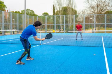 İki genç adam aktif olarak bir pikballa oynuyorlar, biri sarı topa kürekle vuruyor, açık hava sahasında, boş zamanlarının ve formundaki...
