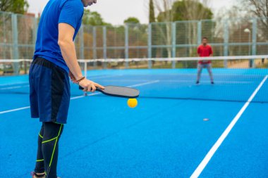 Pickleball müsabakasında oynayan oyuncu açık hava sahasında, arka planda rakibi olan siyah bir kürekle sarı topa vurmaya hazır.