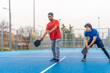 İki genç adam açık hava sahasında yankesicilik oynuyor, biri sarı topa kürekle vurmak üzere, arkadaşı izlerken, enerjik ve rekabetçi bir eğlence.