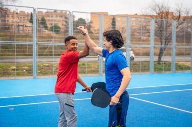 İki gülümseyen erkek sporcu, bir Afro-Amerikalı ve bir Kafkasyalı, mavi bir açık hava pikselbol sahasında beşlik çakıyor, takım çalışmasını ve maçtan sonra iyi bir sportmenliği kutluyor.