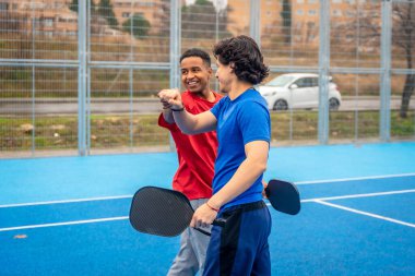 İki farklı erkek sporcu, bir oyun ya da antrenman sonrası işbirliği, yoldaşlık ve takım ruhunu simgeleyen açık hava mavi bir picleball sahasına yumruk atıyor.