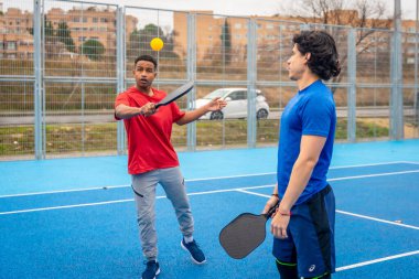 Pickleball oynamaktan hoşlanan genç erkekler, canlı bir mavi sahada topa raketle vurmak, açık havada aktif bir yaşam tarzı ve arkadaşlığı somutlaştırmak.