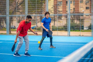 İki farklı genç adam, güneşli kentsel gökyüzünün altında birlikte spor ve zindeliğin keyfini çıkarırken, odaklanmış ve enerjik bir şekilde mavi bir pikbol sahasında yarışıyor.