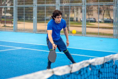 Mavi bir sahada, elinde raketini tutarak topa vurmaya hazırlanan genç bir adam. Ağa odaklanmış ve atletik bir şekilde.