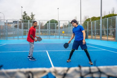 Pickleball oynayan iki genç adam, açık hava mavi kortunda oynanan rekabetçi bir maçta topa vurarak, odaklanma ve aktif yaşam tarzı gösteriyorlar.