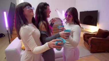 Moda gözlüklü, neşeli bir ev partisinde eğlenen üç genç kadın ellerinde fincanlarla dans ediyor ve neon ışıklarla aydınlanan modern bir oturma odasında kutluyorlar.