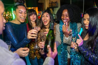 Özel bir olayı kutlarken gülümseyen ve gülen bir grup heyecanlı yetişkin arkadaş, canlı bir partide bir şişe şampanya ve köpüklü içeceklerin tadını çıkarıyorlar.