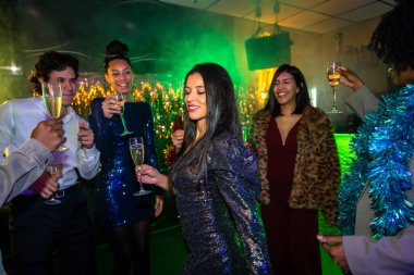 Çeşitli yetişkin arkadaşlar dans ediyor ve şampanya bardaklarını tokuşturuyor. Neşeli bir gecenin tadını çıkarıyorlar. Aydınlatma, kutlama, neşe ve beraberliği yansıtıyorlar.