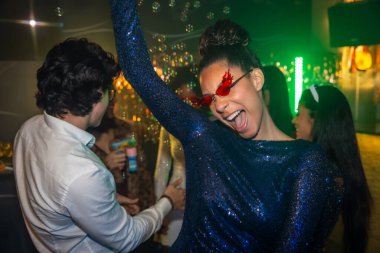 Kırmızı gözlüklü, pullu elbiseli genç bir kadın hareketli bir gece kulübünde arkadaşlarıyla dans edip kutlama yapıyor. Etrafında kabarcıklar uçuşuyor. Enerji, neşe ve canlı bir atmosfer yayıyor.