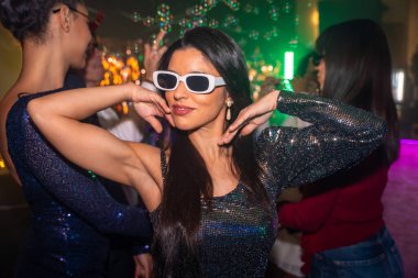 Göz alıcı pullu elbisesi ve şık beyaz güneş gözlüklü genç bir kadın neşeli bir gece atmosferinde arkadaşlarıyla kutlama yaparak şen şakrak bir disko partisinin tadını çıkarıyor.