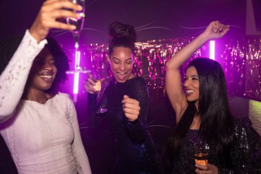 Mor ışıklar altında canlı bir gece kulübünde dans eden ve tezahürat yapan çeşitli genç kadınlar. Birlikte neşeli bir gece geçirip gülümsüyorlar.