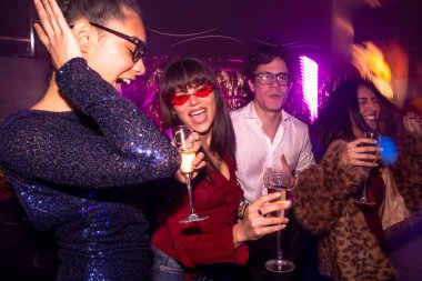 Bir grup farklı arkadaş özel bir olayı kutluyor, şampanya içiyor, dans ediyor ve mor ışıklarla dolu canlı bir gece kulübünde eğleniyorlar.