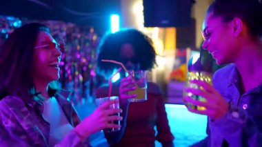 Üç mutlu çoklu etnik kadın bardaklarını tokuşturuyor, kokteyller içiyor ve canlı bir gece kulübünde birlikte dans ediyor, arkadaşlıklarının tadını çıkarıyor ve renkli neon ışıkları altında kutlama yapıyorlar.