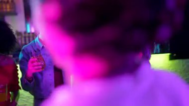 Çok ırklı bir arkadaş grubunun güldüğü, kokteyl içtiği ve bir gece kulübünde dans ettiği parlak mor ve mavi neon ışıkların altında fütürist parlak gözlüklerle kutlama yaptığı canlı bir sahne.