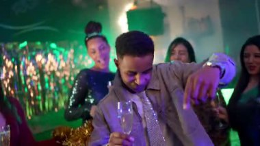 Arkadaşlarıyla dans eden ve elinde bir bardak şampanya tutan mutlu genç adam renkli neon ışıkları ve şenlikli süslemeleri olan bir gece kulübündeki partinin canlı atmosferinin tadını çıkarıyor.