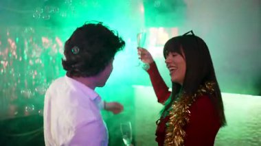 Çeşitli gülen arkadaşlar bir gece kulübünde şampanya bardaklarıyla dans edip kadeh kaldırıyorlar. Neşeli insanlar renkli disko ışıkları altında alkol ve baloncuklarla kutlama yapıyorlar.