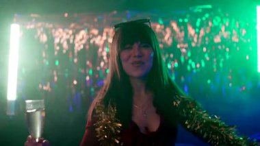 Mutlu genç bir kadın kameraya dans ediyor ve gülümsüyor Noel arifesinde ya da yeni yıl partisi kutlamalarında, elinde bir bardak şampanya tutarken ve etrafı süs ve neon ışıklarıyla çevriliyken