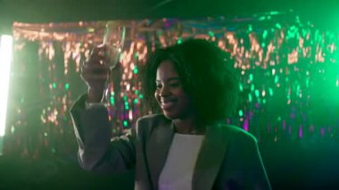 Bir partide kıvırcık saçlı, dans eden ve şampanya içen neşeli genç bir kadın. Parlak, şenlikli bir arka planda, bir gece kulübünün canlı yeşil ve mor ışıklarında mutlu bir şekilde gülümsüyor.