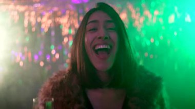 Gece kulübünde dans eden kürklü çekici genç bir kadın elinde bir bardak şampanya, renkli ışıklar ve dumanla parti atmosferinin tadını çıkarıyor.