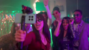 Genç ve şık bir kadın arkadaşlarıyla canlı bir gece kulübünde dans edip şarkı söylerken bir video kaydediyor, canlı atmosferi yakalıyor ve sosyal medya içeriği yaratıyor.