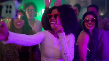 Kırmızı kalp takan genç Afrikalı Amerikalı bir kadın. Güneş gözlüğü takıyor. Bir gece kulübünün neon ışıkları altında arkadaşlarıyla dans ederken akıllı telefonuyla selfie çekiyor.